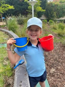 young school student, smiling and holding a blue bucket and a red bucket and wearing a blue St Catherine's School cap